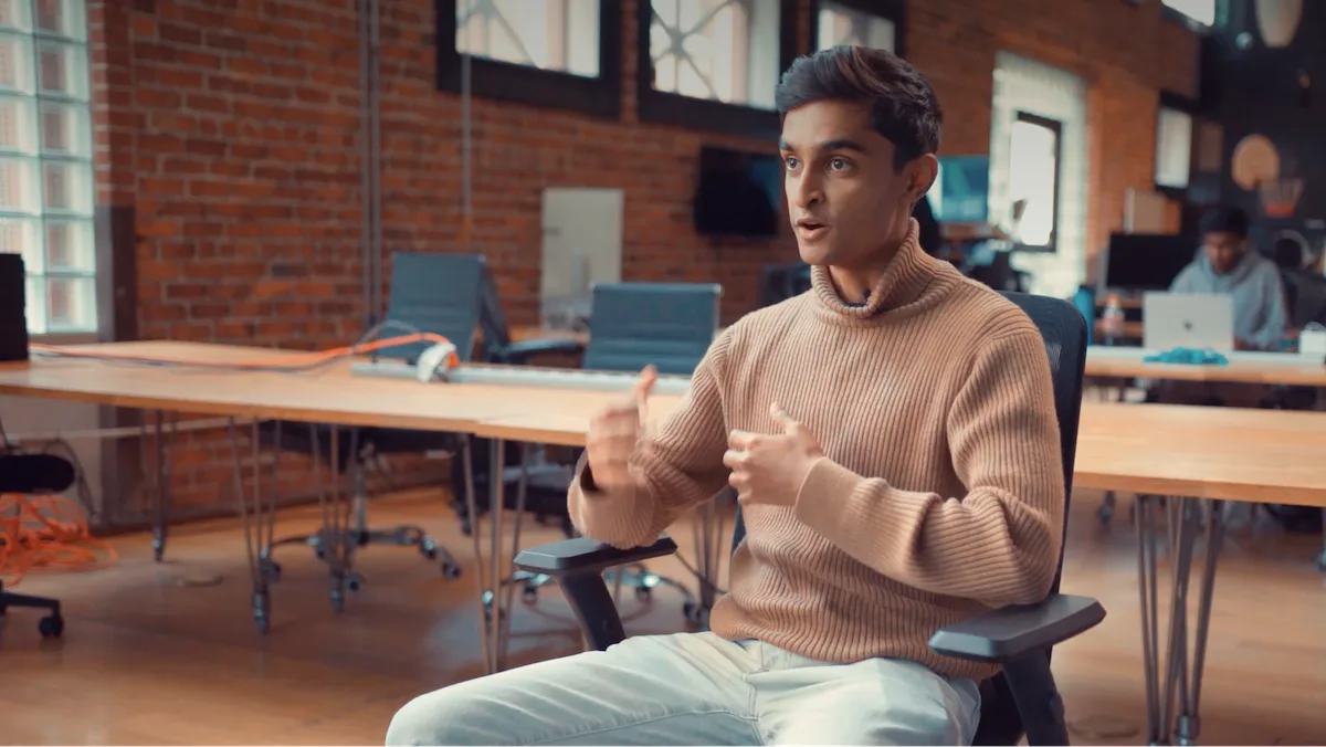 Person sitting in an office chair, wearing a tan sweater, gesturing with hands, in a brick-walled workspace with tables and chairs in the background.